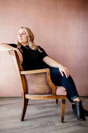 Woman in black outfit posing dramatically on vintage cane chair against pink wall.