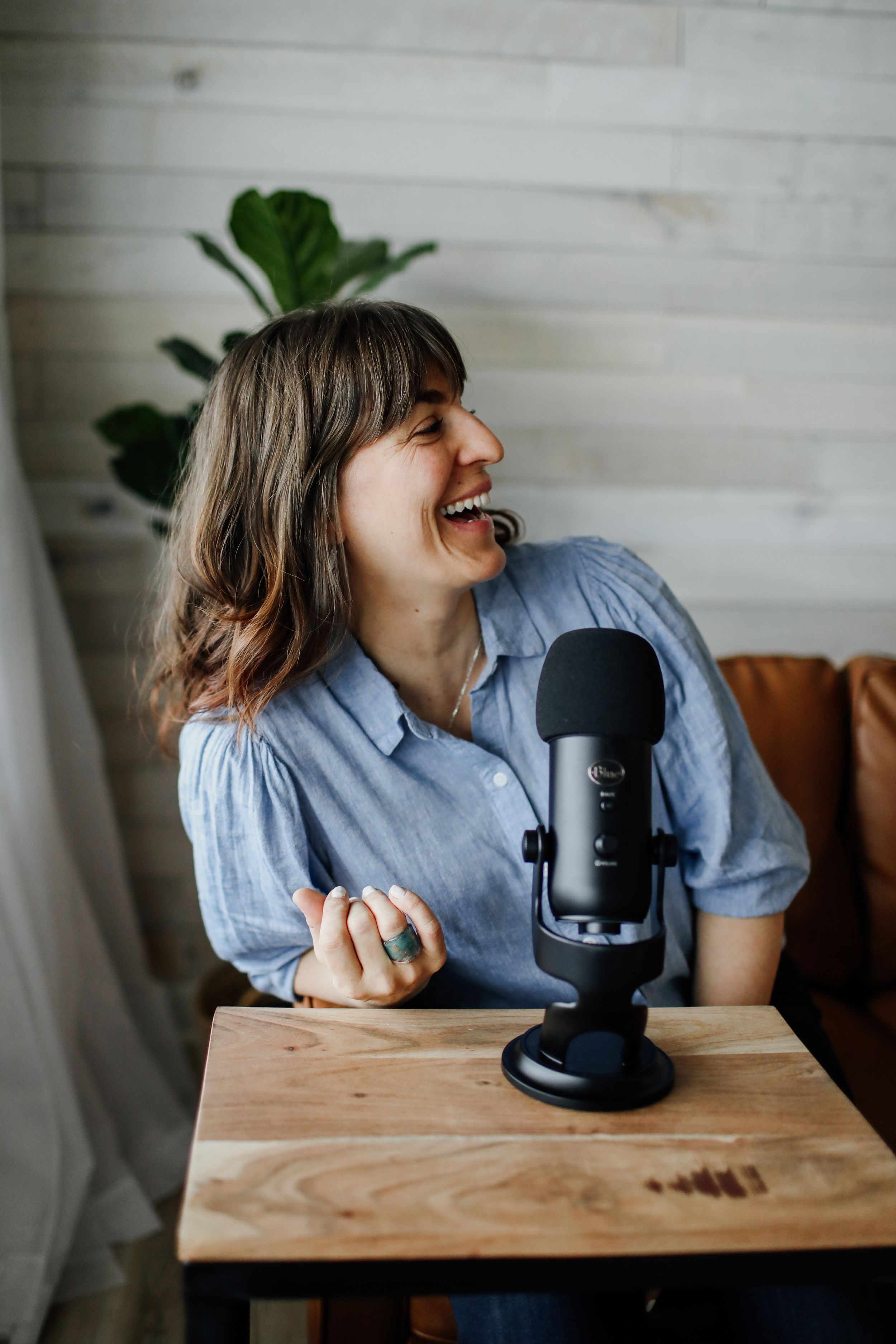 Person laughing while recording podcast with blue microphone on wooden desk.