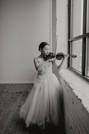 Violinist in an elegant white tulle gown performs by a window in black and white.