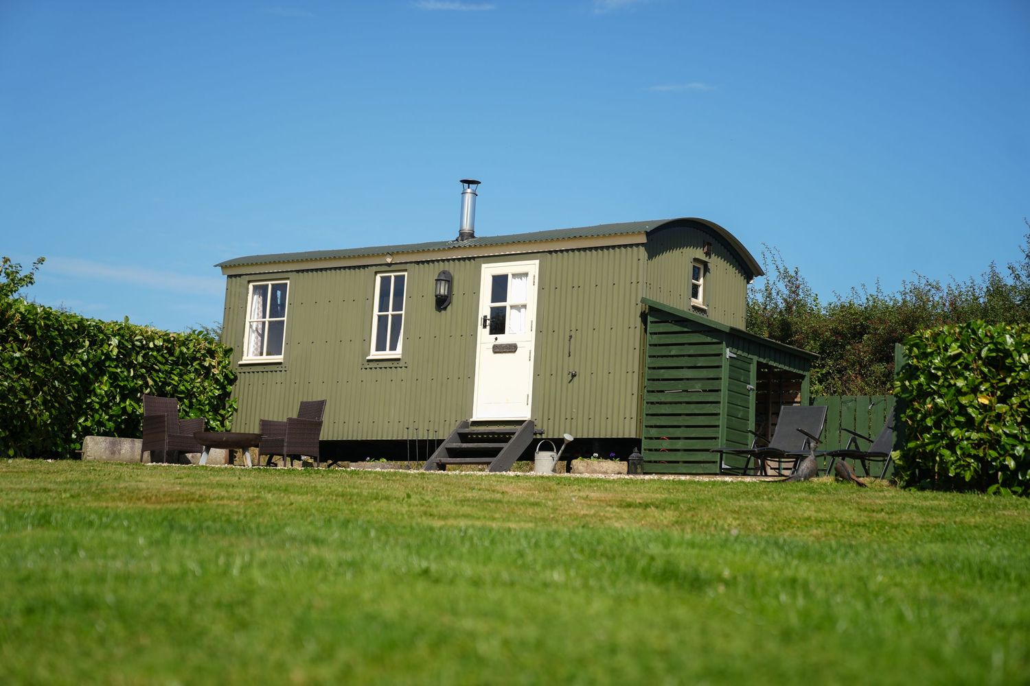 Green shepherd's hut sits on manicured lawn with hedges.