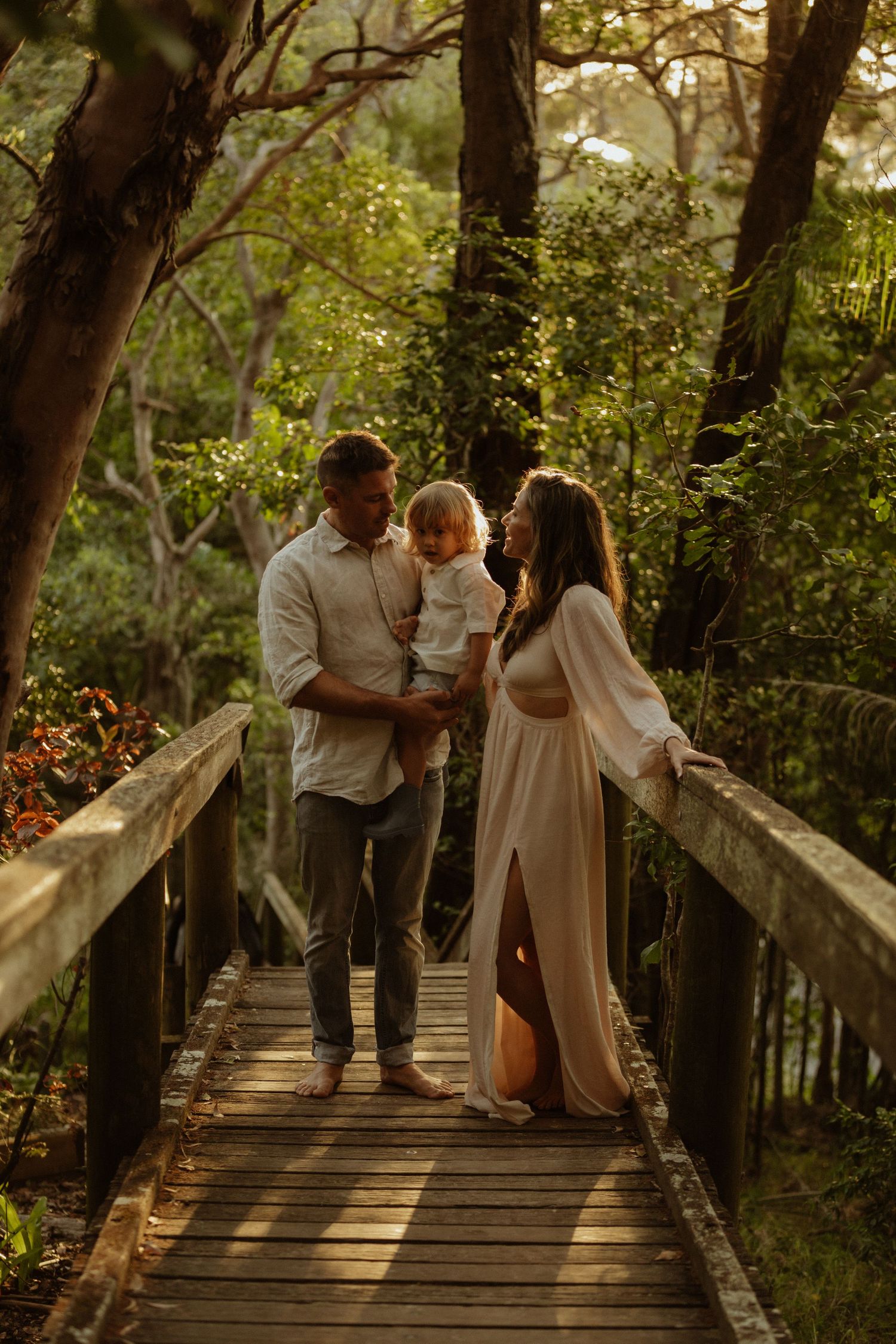 Family stands together on wooden bridge path in sunlit forest setting.