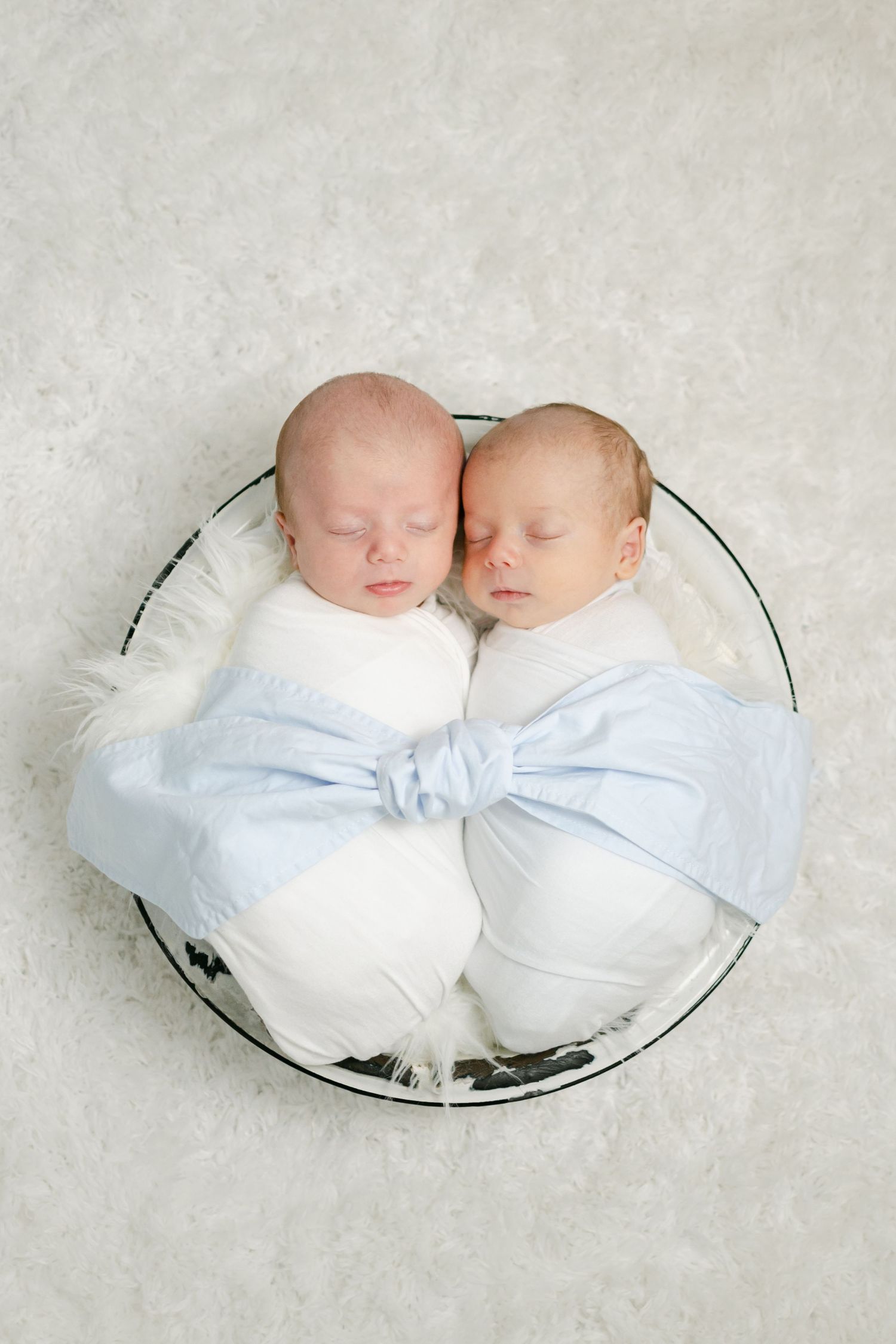 Twin newborns swaddled in white and blue wraps sleeping together in a round basket on white fur.