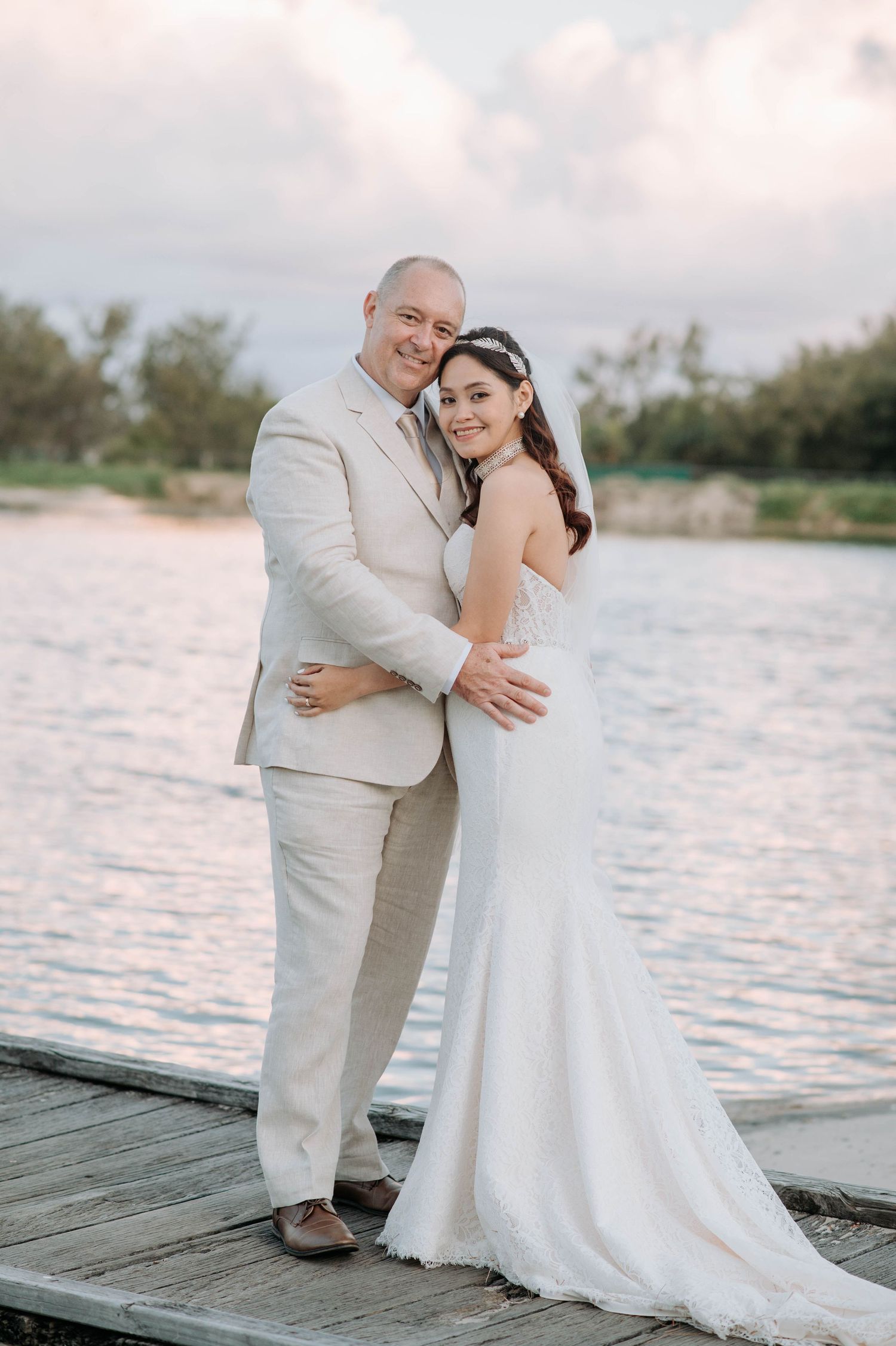 Romantic wedding couple embracing on a waterfront dock at sunset in light colored attire.