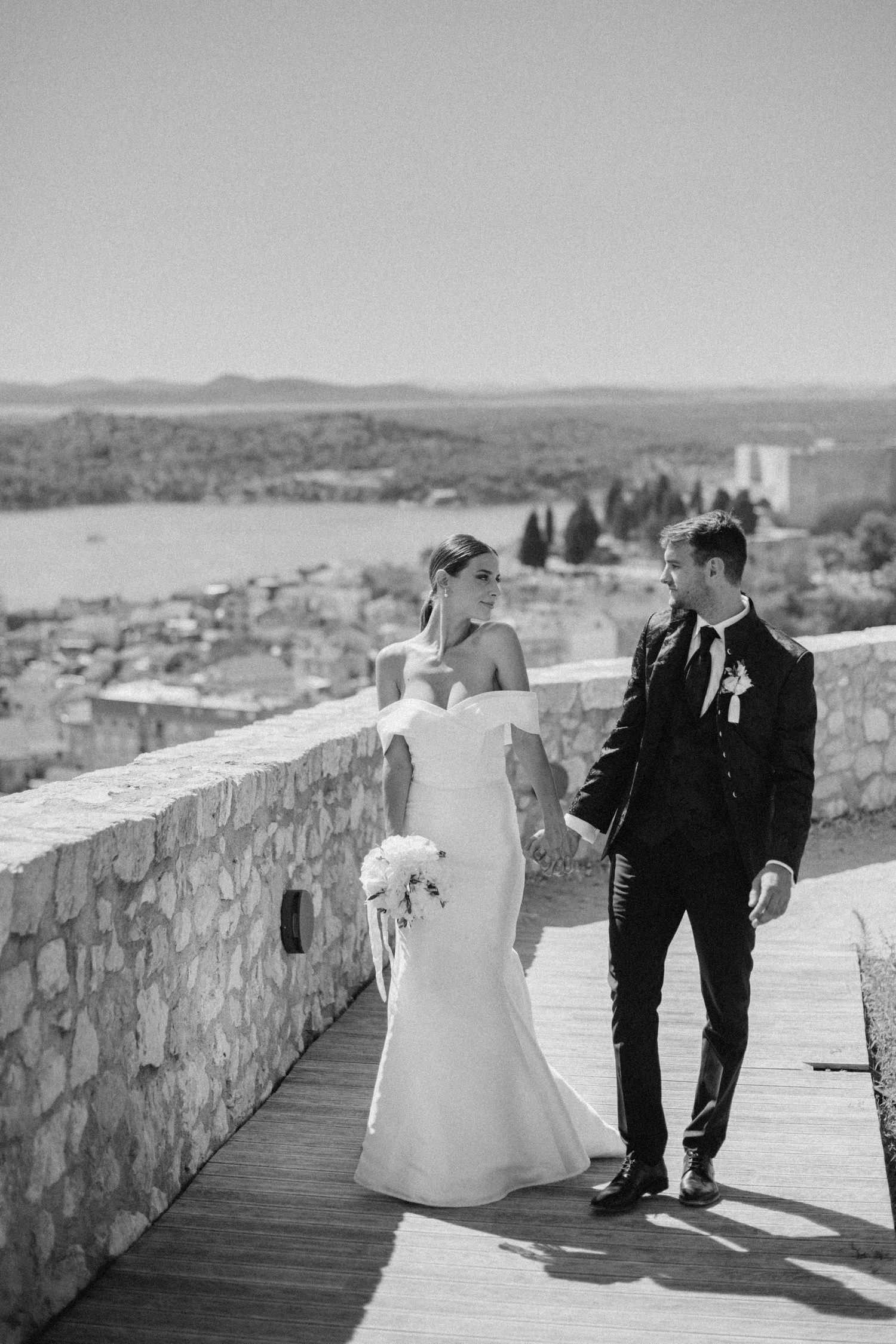 A couple in wedding attire walks along a stone wall overlooking a scenic coastal town on their special day.