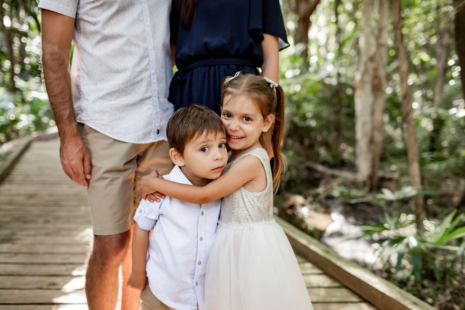 Young siblings embrace while walking on a wooden boardwalk path through lush forest scenery.
