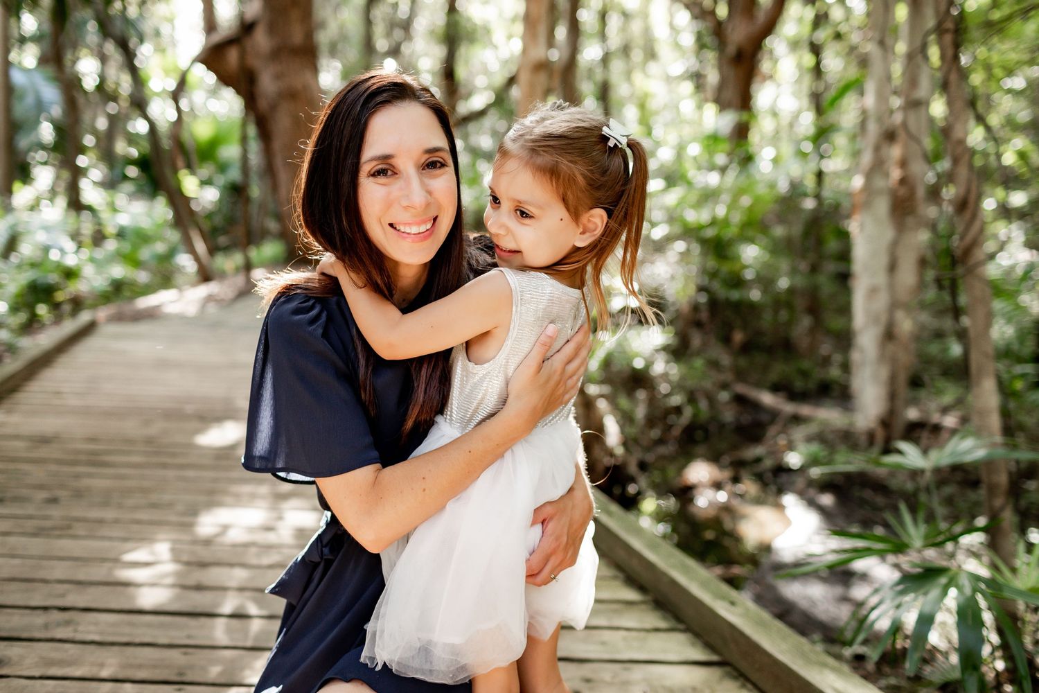 A tender embrace shared between parent and child on a sunlit wooden walkway in a forest setting.