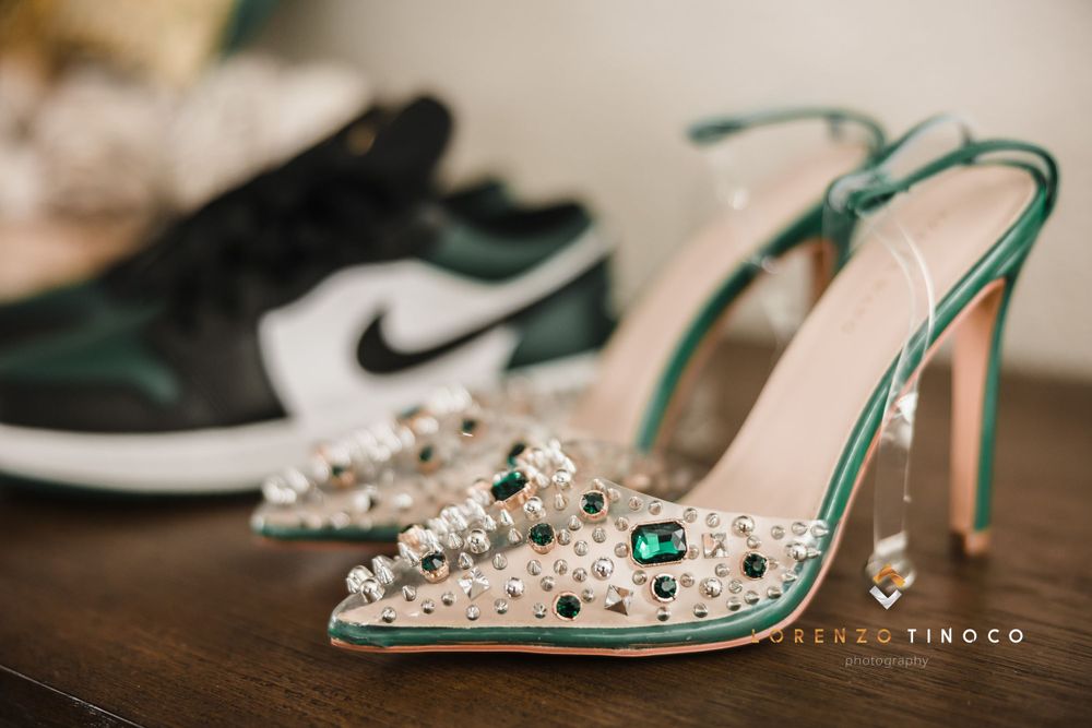 Crystal-embellished clear heels with green accents next to Nike sneakers on wooden surface.