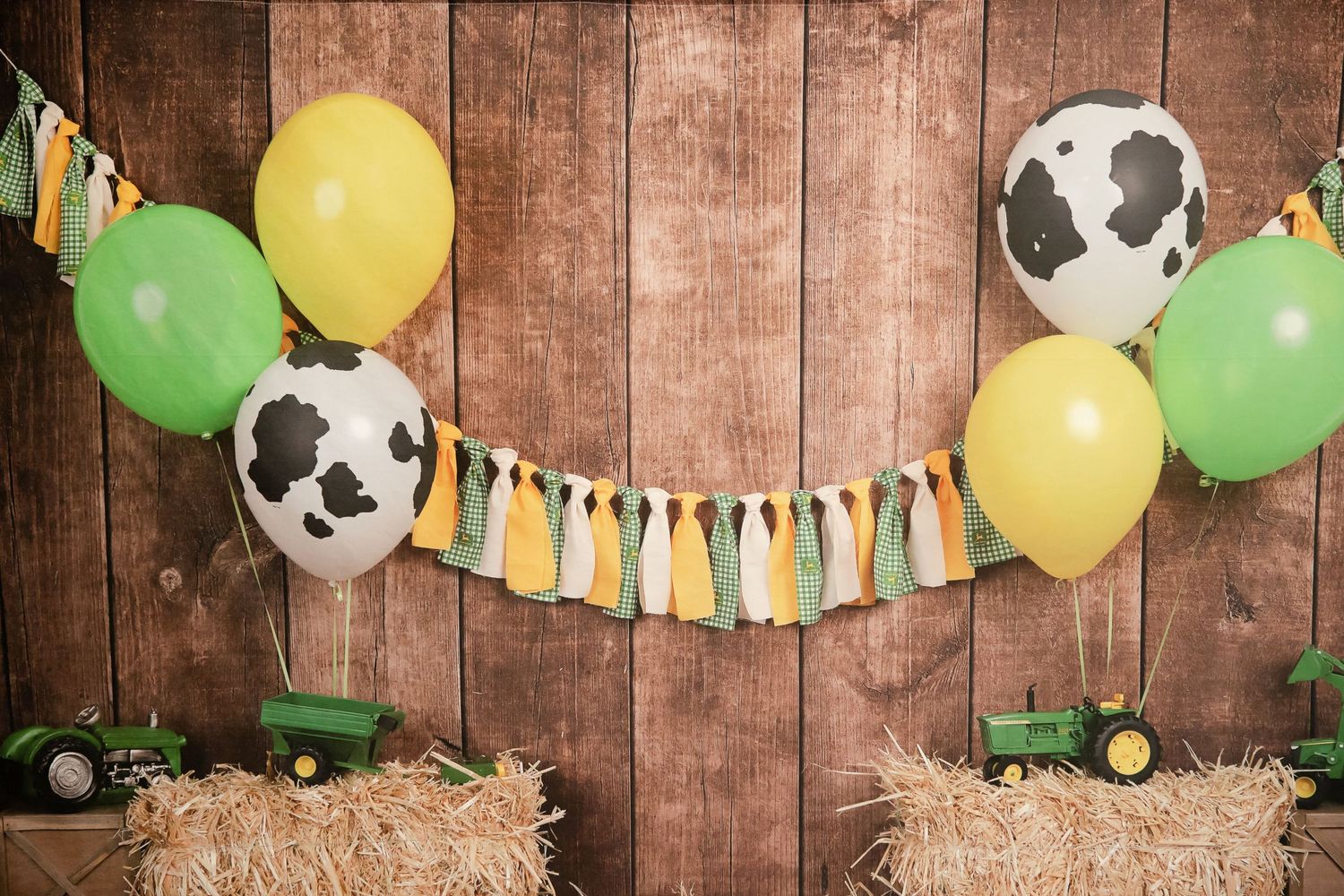 Farm themed party backdrop with cow print balloons, green and yellow bunting, and hay bales.