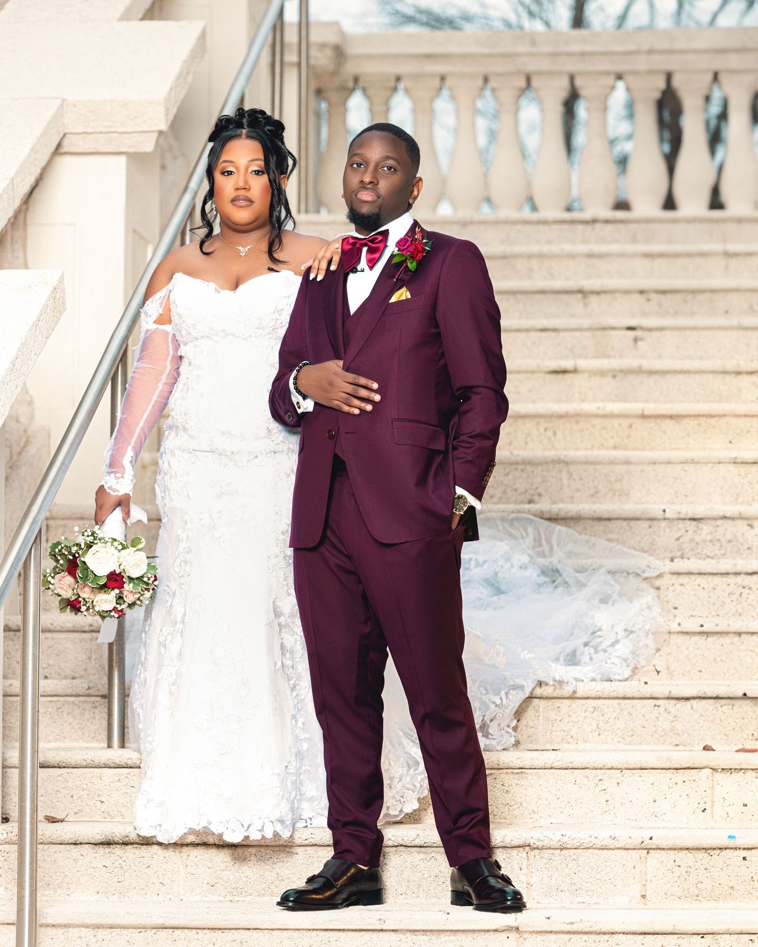 A wedding couple in white dress and burgundy suit posing on marble stairs.