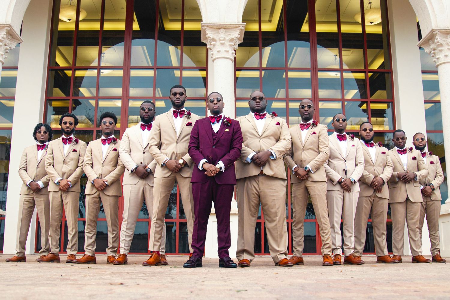 Large wedding party in beige suits with one person in burgundy standing in front of arched building.