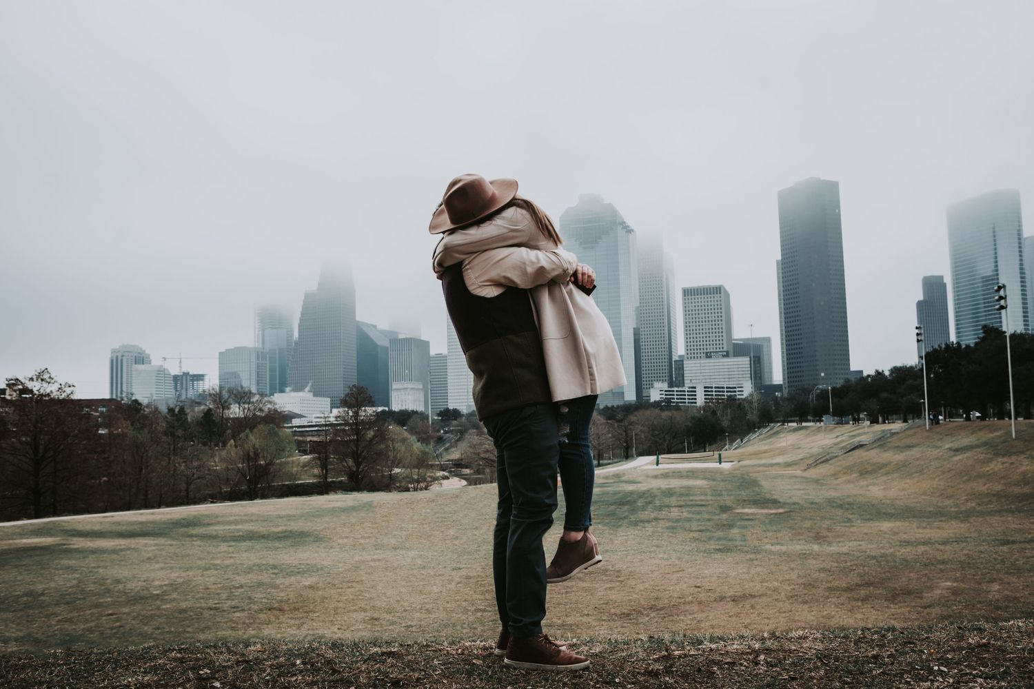 Two people embrace in an intimate moment against a misty urban skyline with skyscrapers in the background.