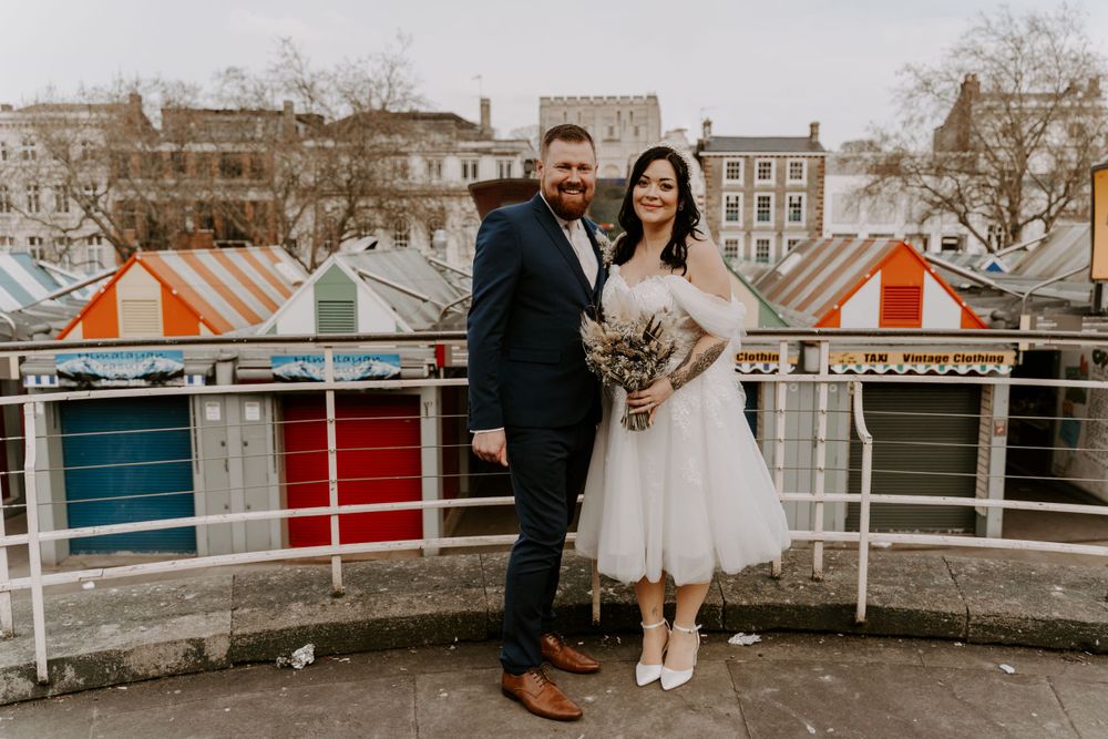 bride and groom standing in the city centre. behind them is a row of colourful market stall roofs.