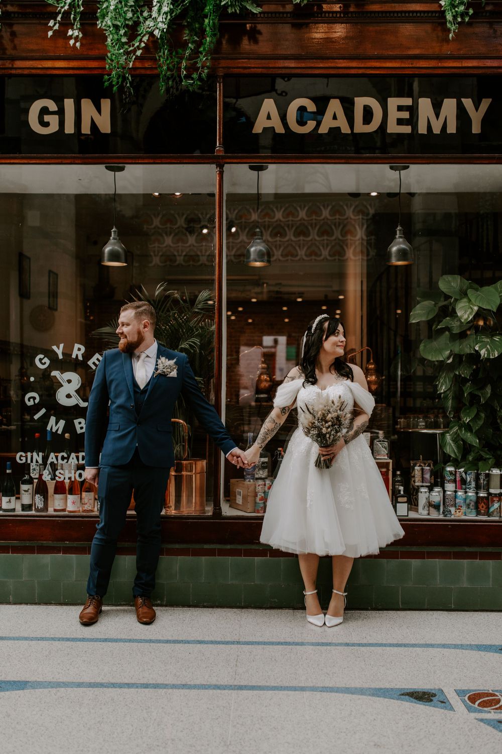 bride and groom standing outside a gin shop in the city centre. they are looking at each other and smiling.