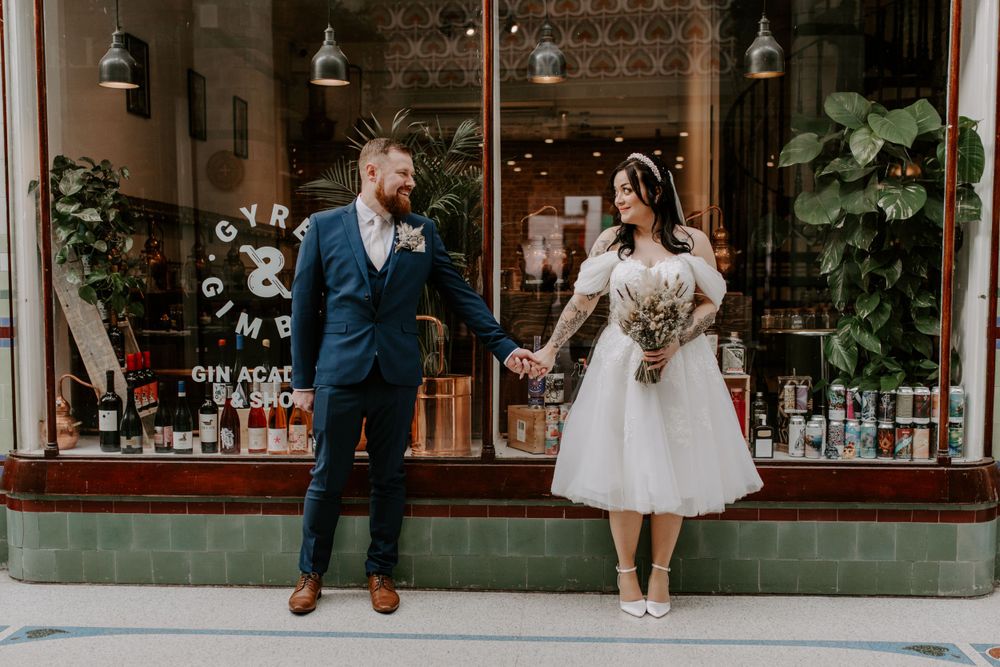 bride and groom standing outside a gin shop in the city centre. they are looking at each other and smiling.
