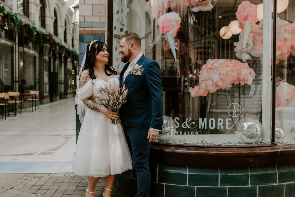 bride and groom standing outside a macaron shop in the city centre. they are looking at each other and smiling.