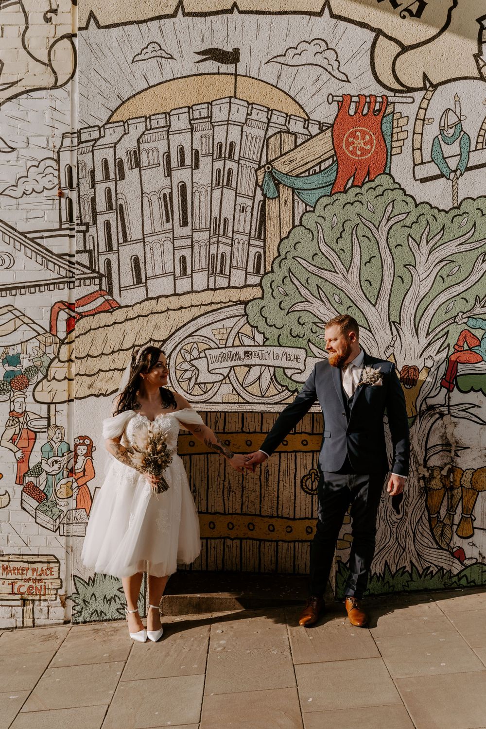 bride and groom standing in front of a graffiti wall holding hands and looking at each other.