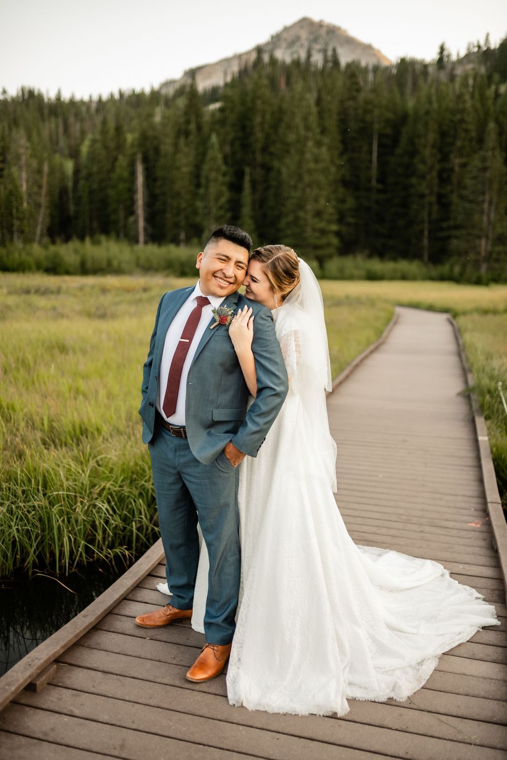 Wedding couple on wooden boardwalk with mountain landscape and pine forest backdrop.
