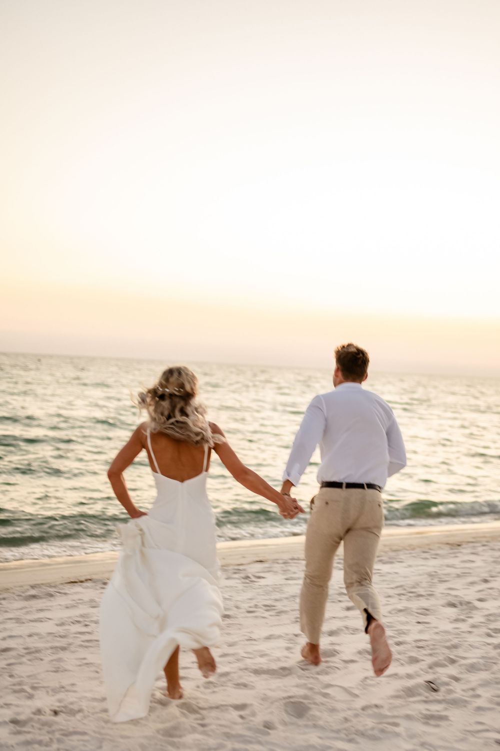 Playful couple in wedding attire jumps together on white sandy beach at sunset.
