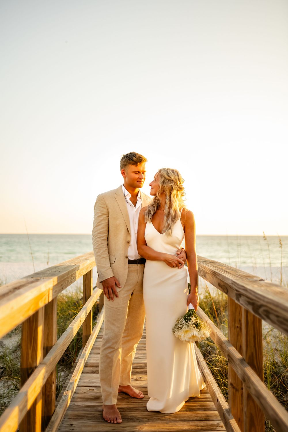 Newlyweds in white and beige attire pose on wooden boardwalk at beach during sunset.