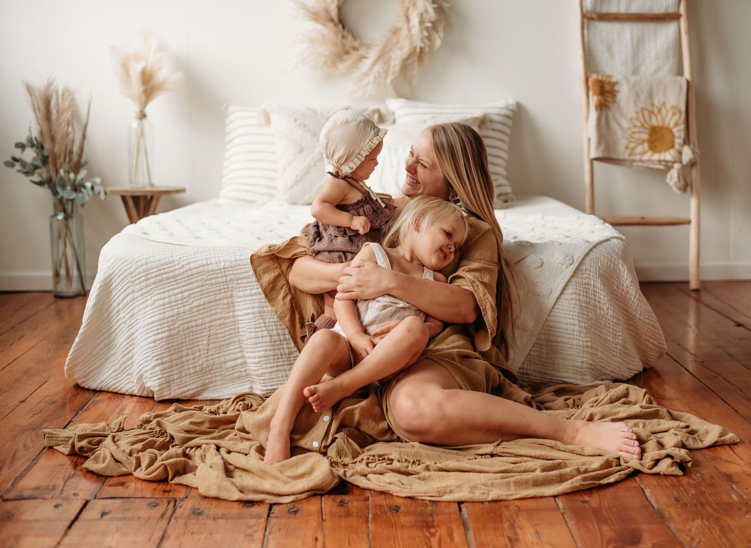 Mother sits on wooden floor holding two young children wrapped in a golden tan blanket.