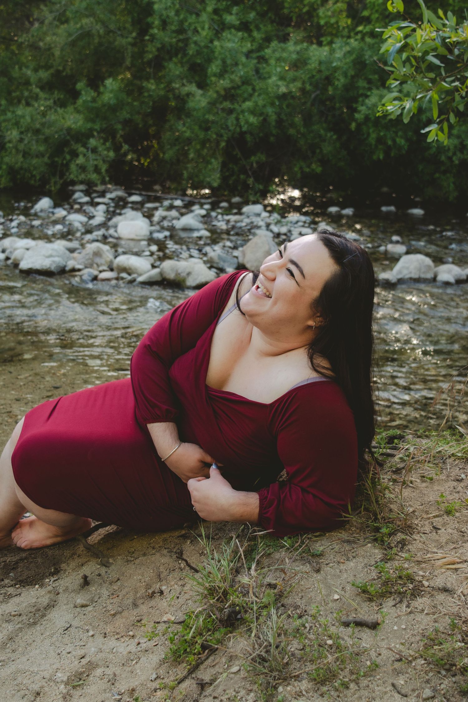 Person in burgundy dress laughing while relaxing by rocky riverside.