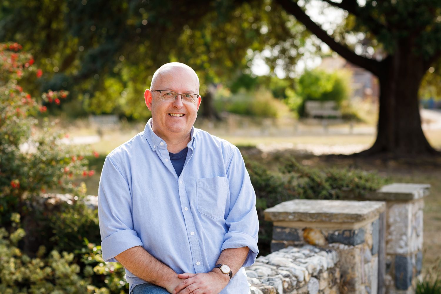 Professional outdoor portrait with person seated on stone wall with garden and trees in background during business branding photoshoot.