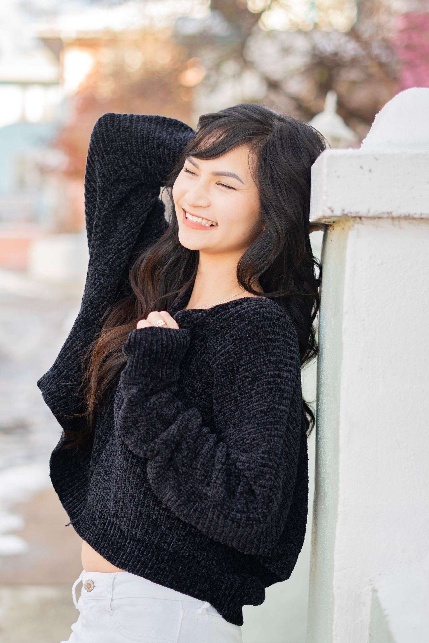 A smiling young woman with long dark hair leans against a white pillar wearing a black knit sweater outdoors.
