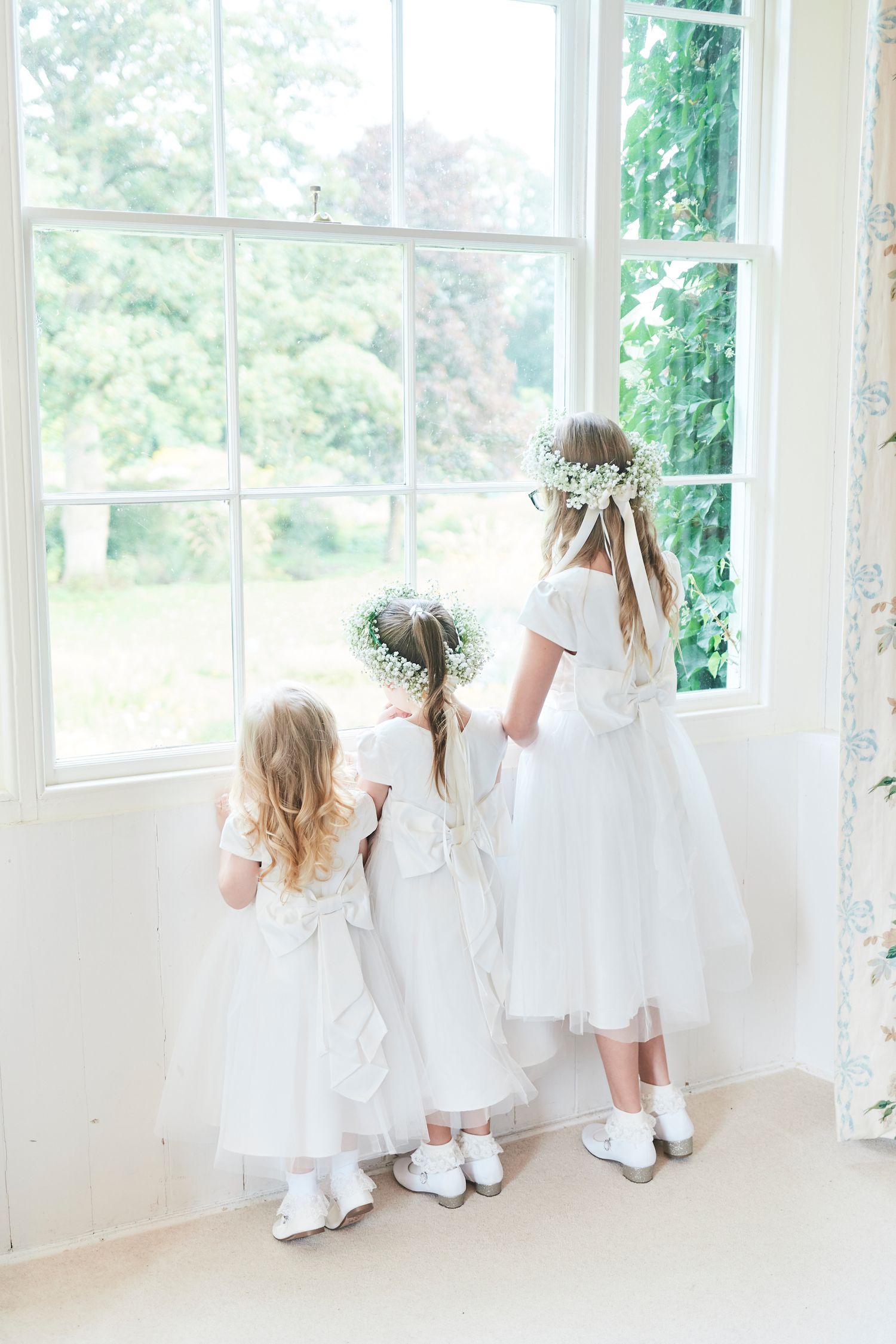 3 flower girls look out of the window in their dresses, at Bressingham Hall, Suffolk. by wedding photographer Faye Amare