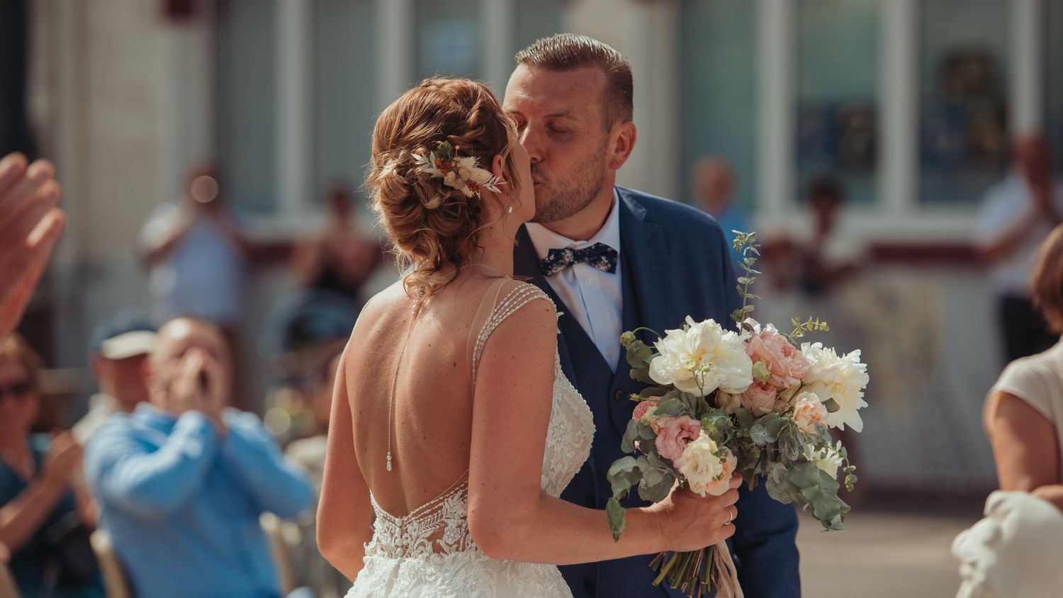 Groom kisses bride on the cheek as she holds a bouquet at an outdoor wedding celebration.