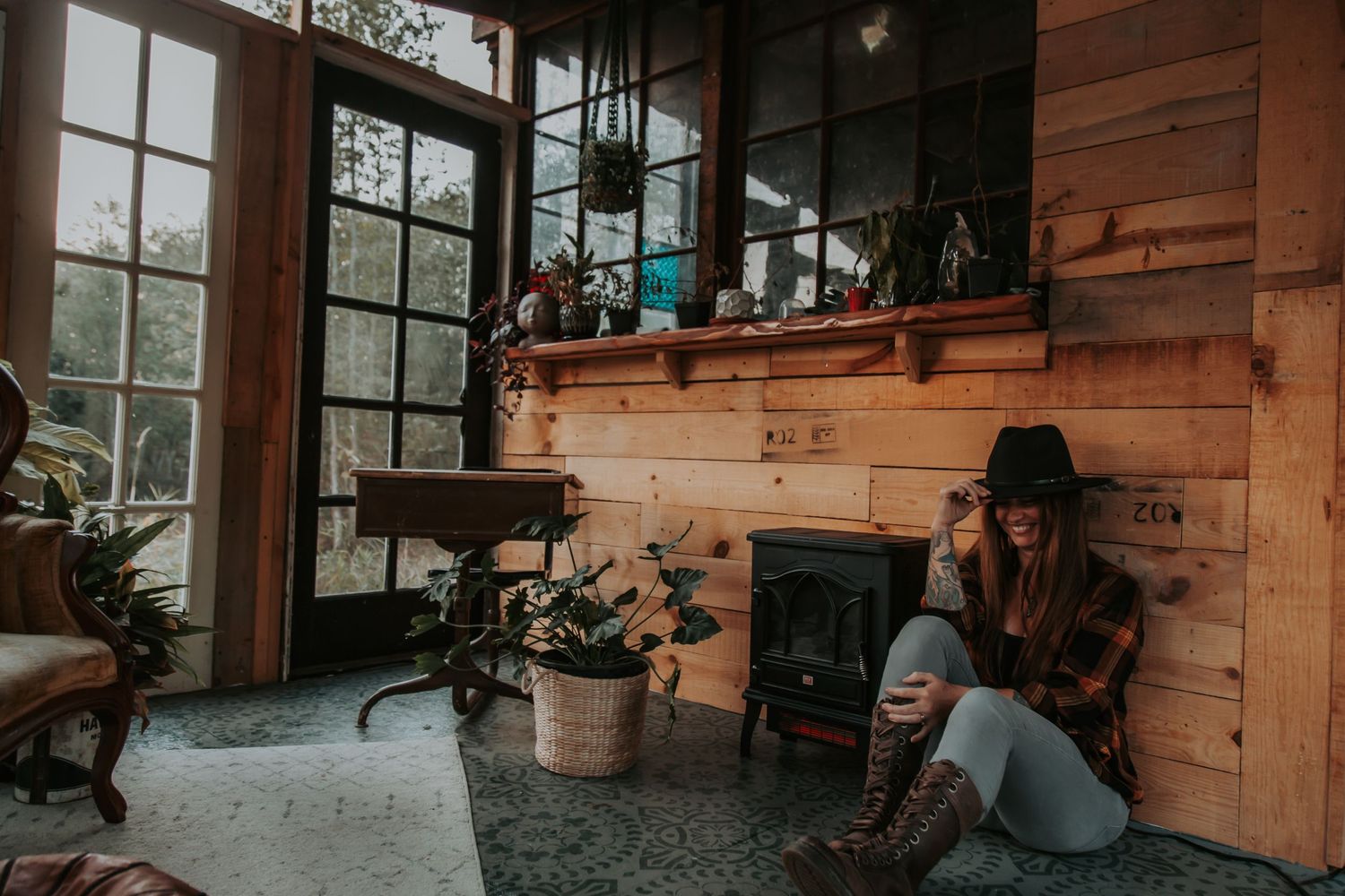Woman in a black hat and plaid shirt sitting by a wood stove in a cozy rustic cabin sunroom filled with plants.