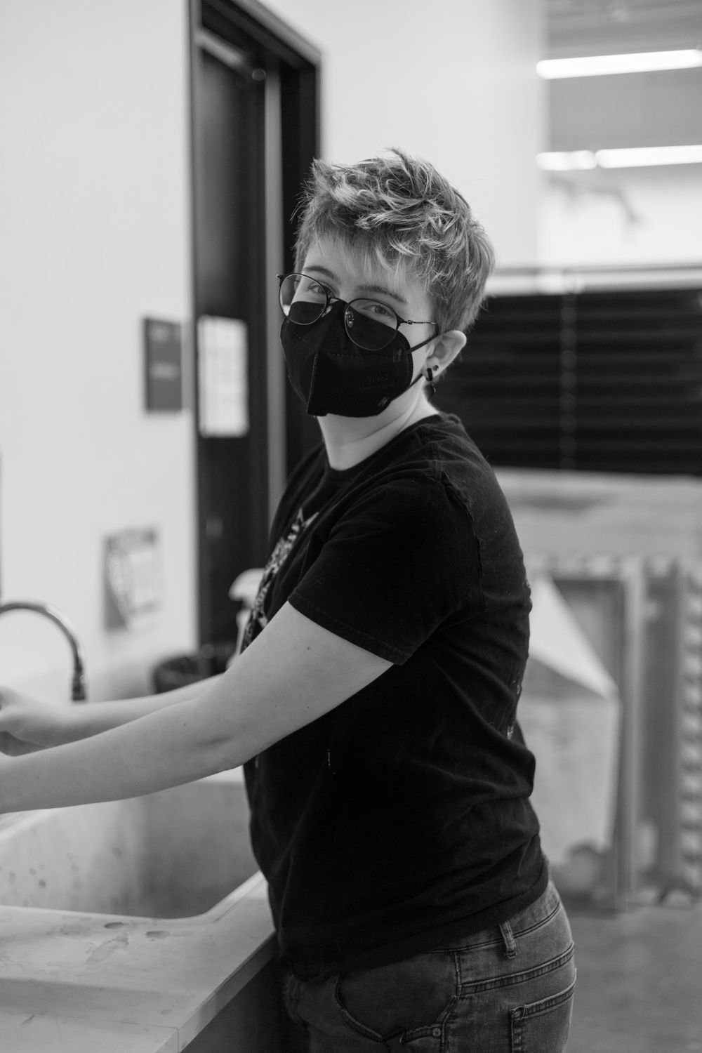 Black and white image of someone wearing a mask and t-shirt while working at a sink.