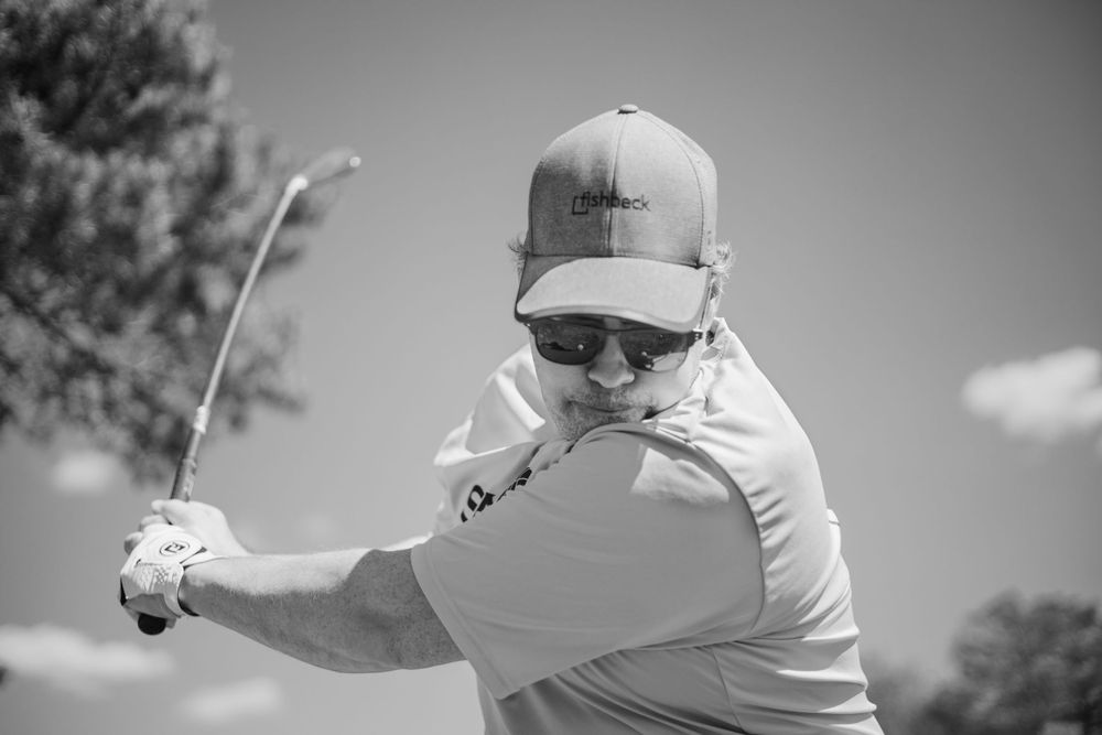 Golfer in mid-swing motion on a sunny day wearing a baseball cap and sunglasses.