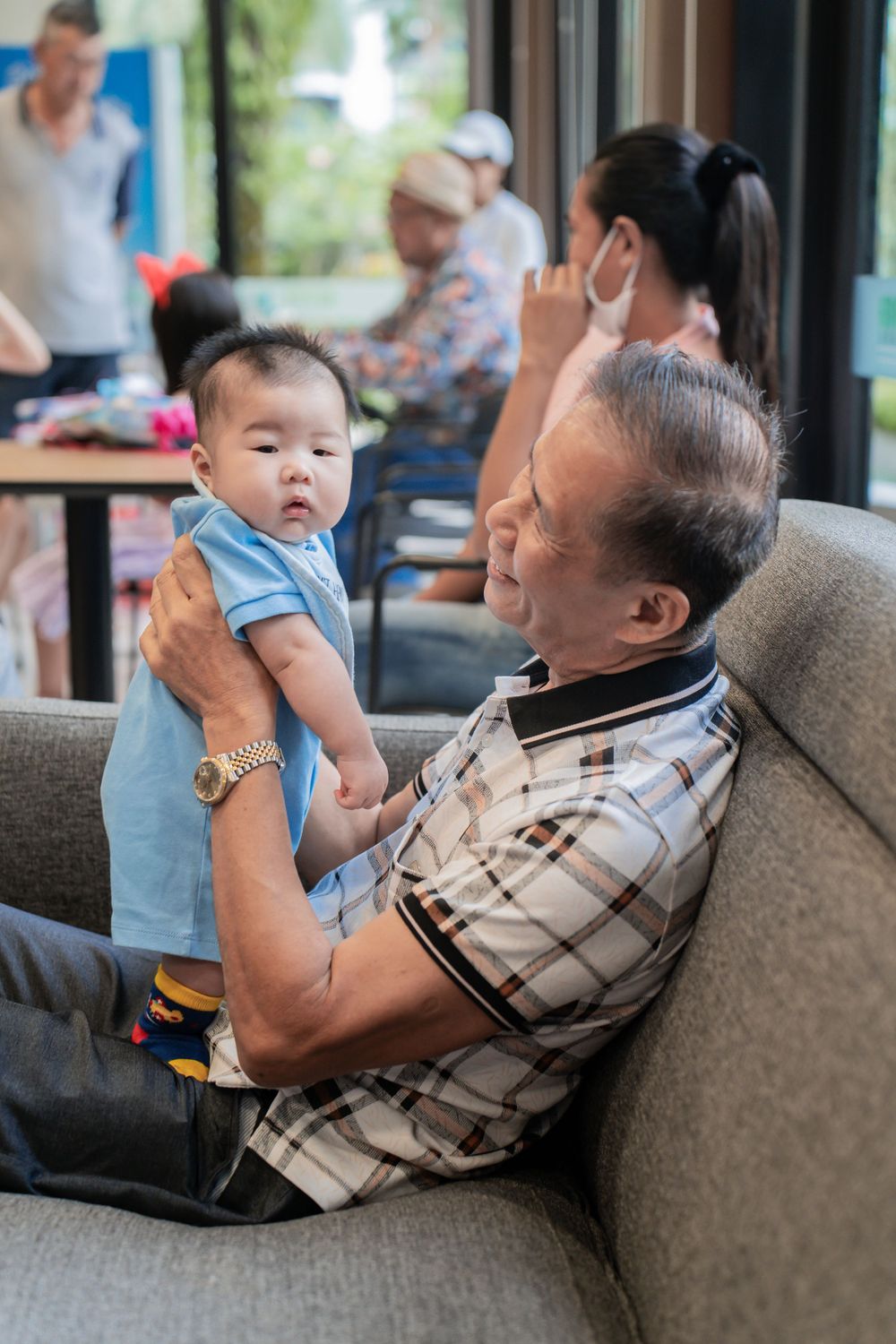A baby in light blue clothing sits with an adult on a gray couch.