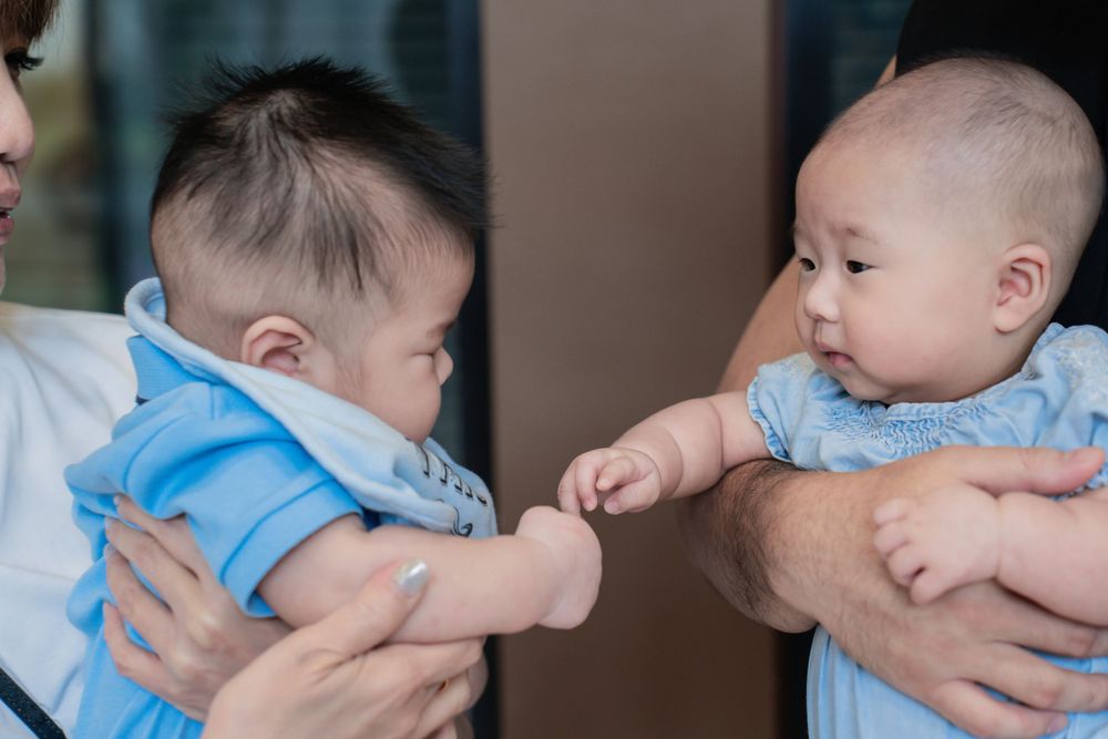 Two babies in matching blue outfits reaching toward each other while being held.