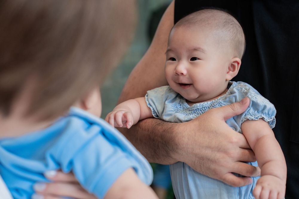 A smiling baby in light blue clothing being held and interacting with others.