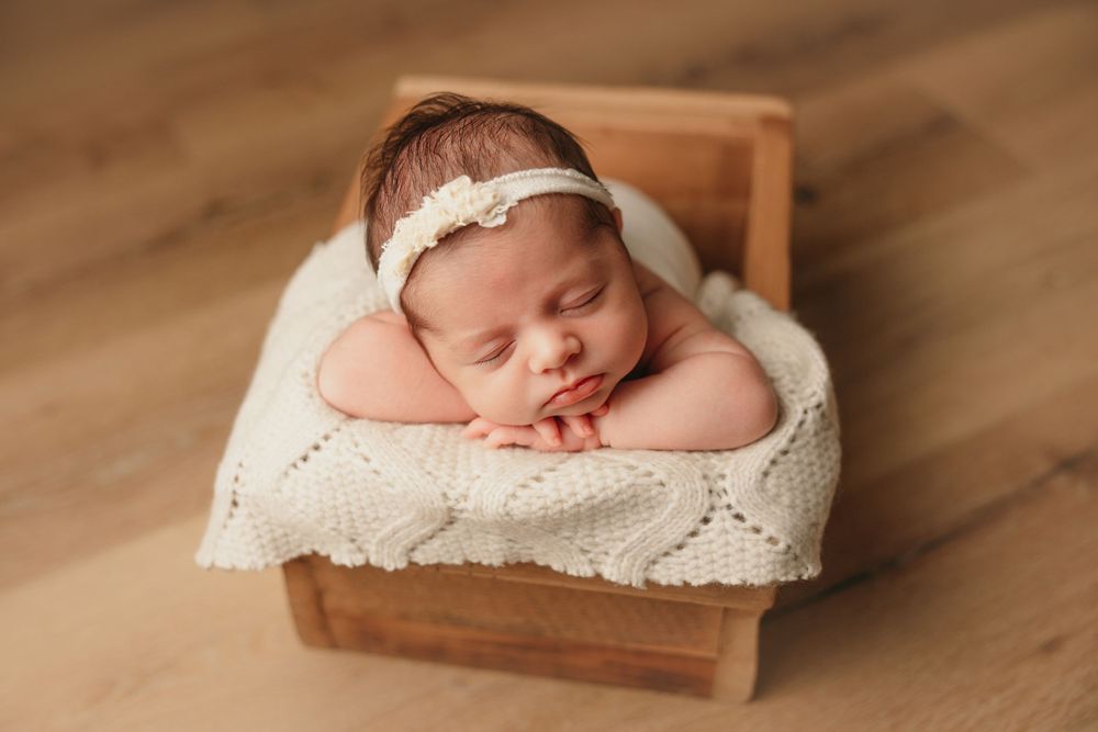 Sleeping newborn in lace headband resting in wooden box with knit blanket.
