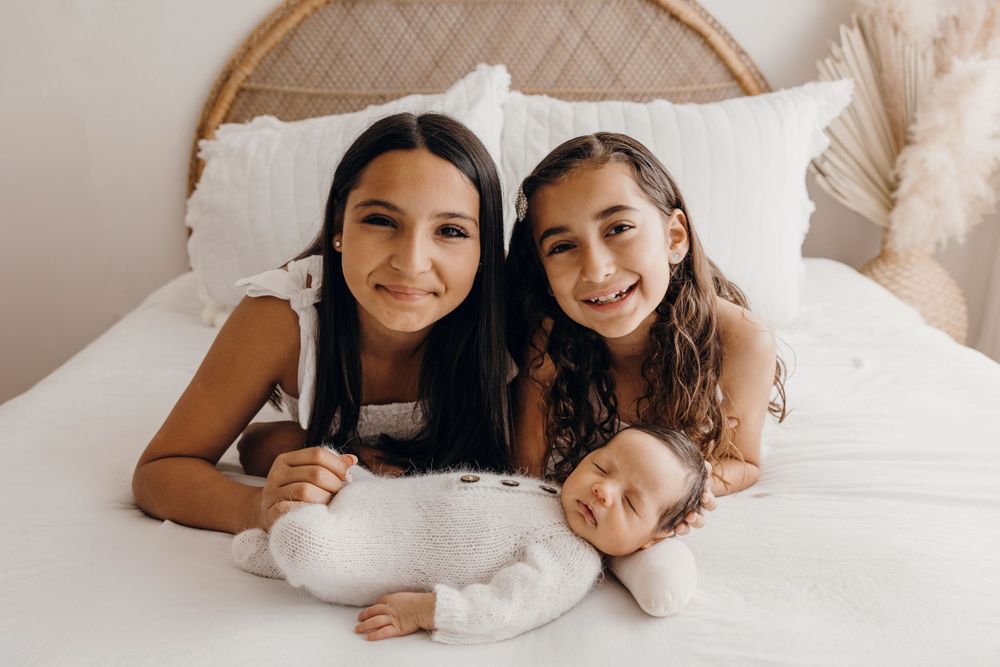 Heartwarming family portrait of siblings cuddling with newborn baby on white bed.