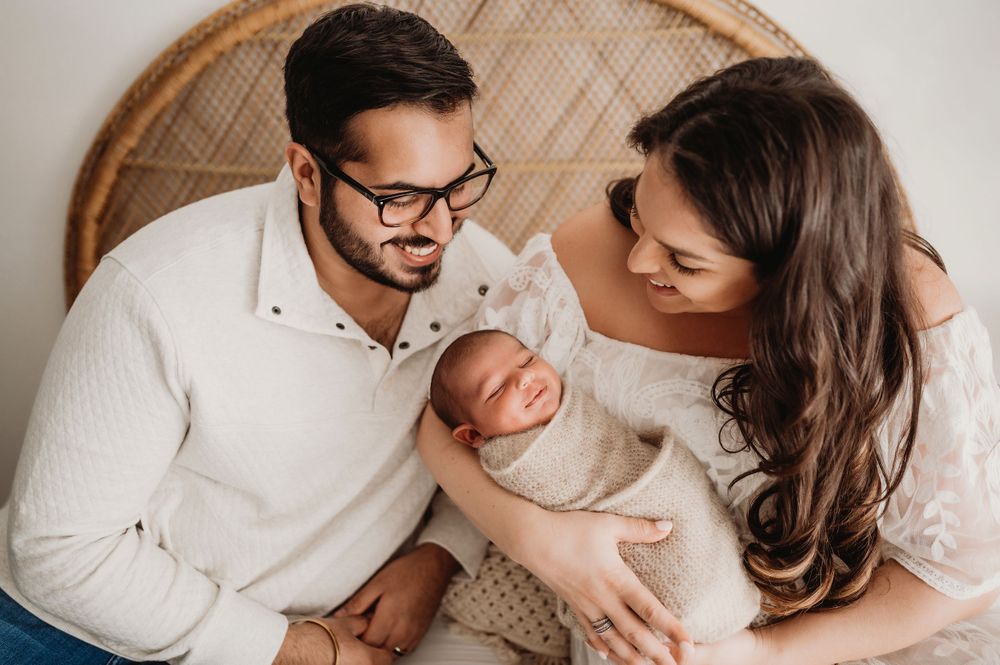 A tender newborn photography session featuring parents in white outfits snuggling with their baby against a woven backdrop.