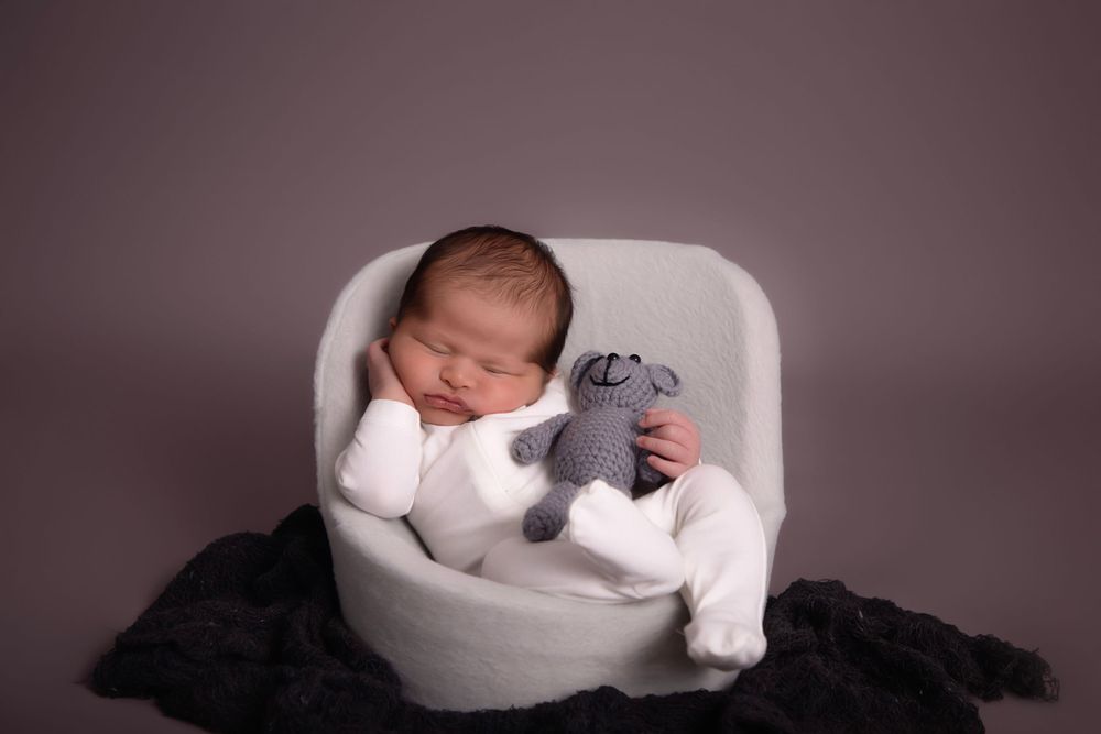 Sleeping newborn in white outfit rests in cozy chair with plush toy.