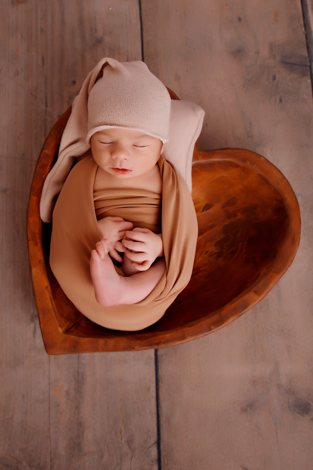 Sleeping newborn wearing beige hat and wrap while resting in wooden heart-shaped bowl.