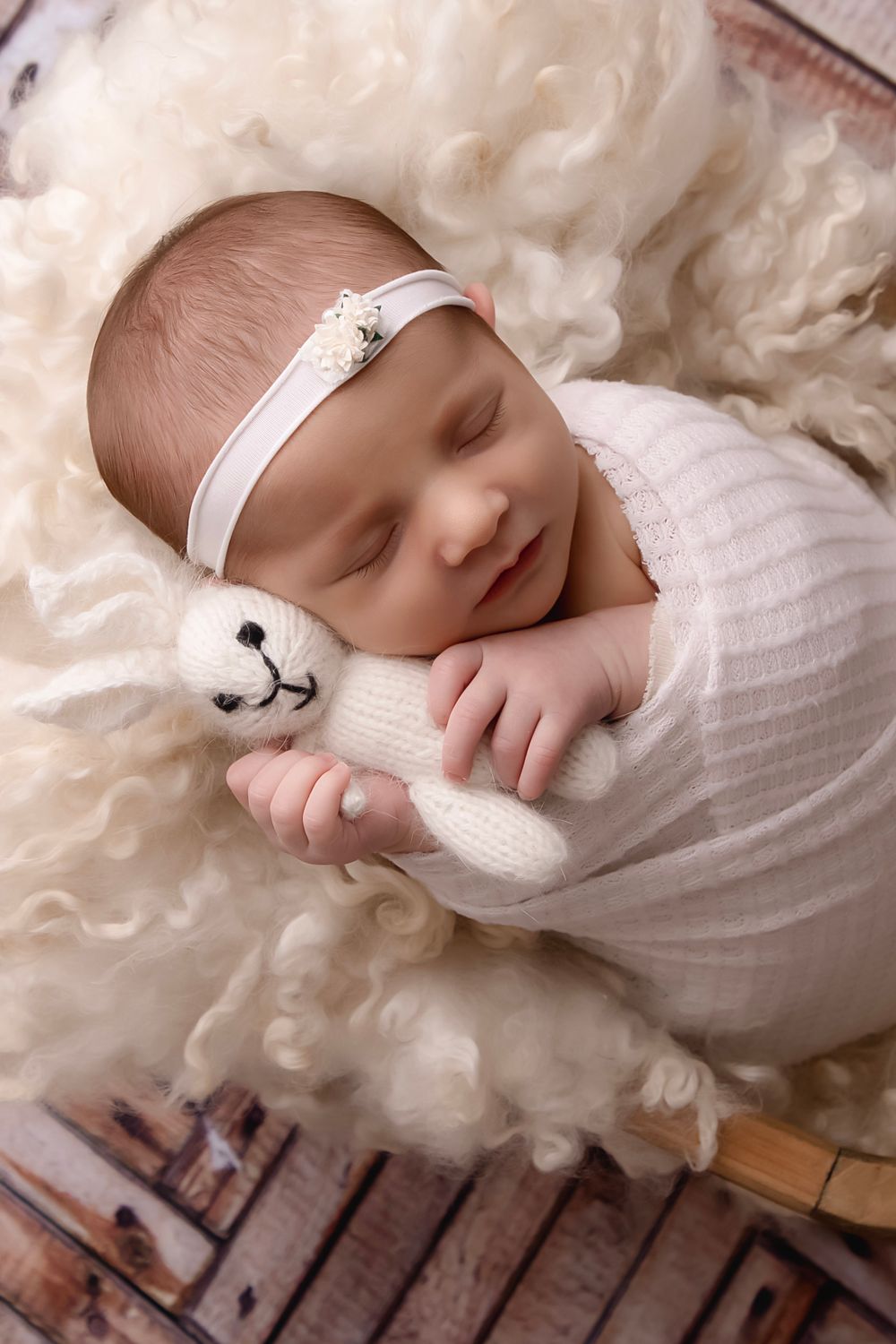 Newborn portrait featuring soft white wrapping and headband against fluffy cream backdrop.