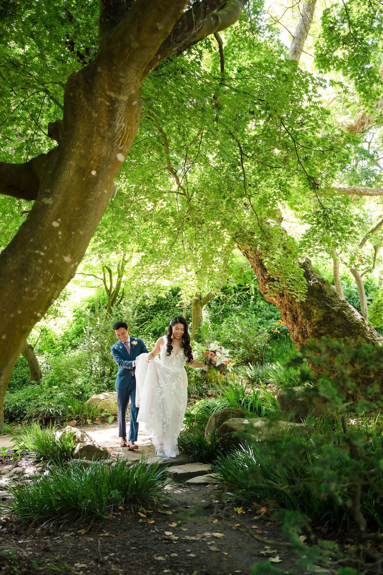 A couple walks along a lush garden path under towering trees with dappled sunlight filtering through the canopy.