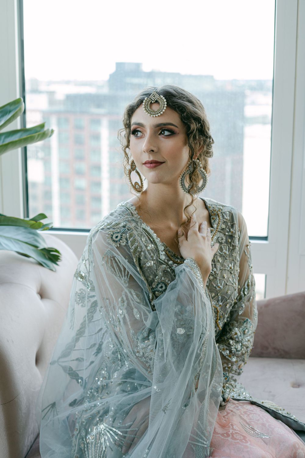 A dreamy bridal portrait by a window with cityscape views wearing an embellished silver ensemble.
