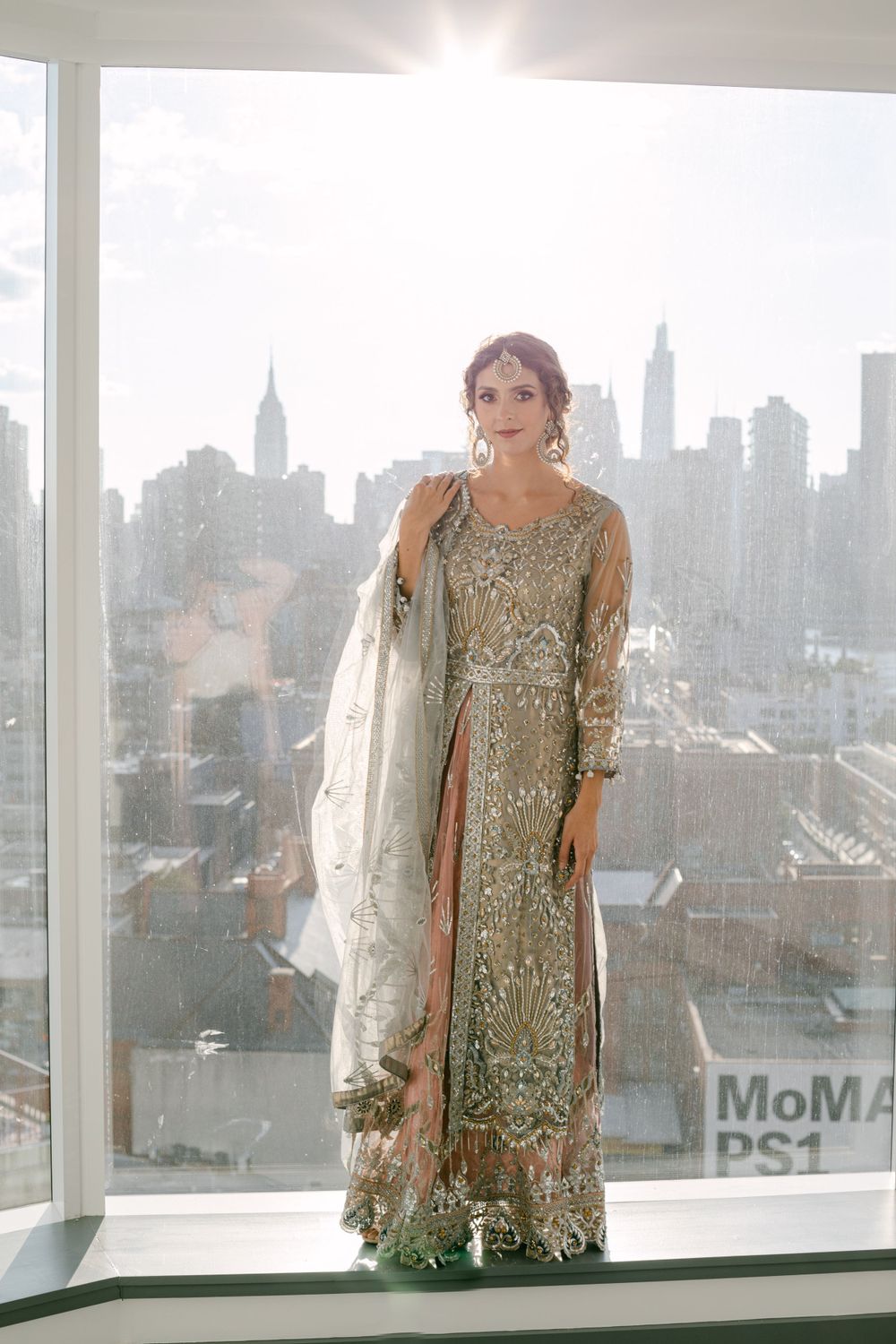 Intricately embellished formal dress photographed against Manhattan skyline with MoMA PS1 visible.
