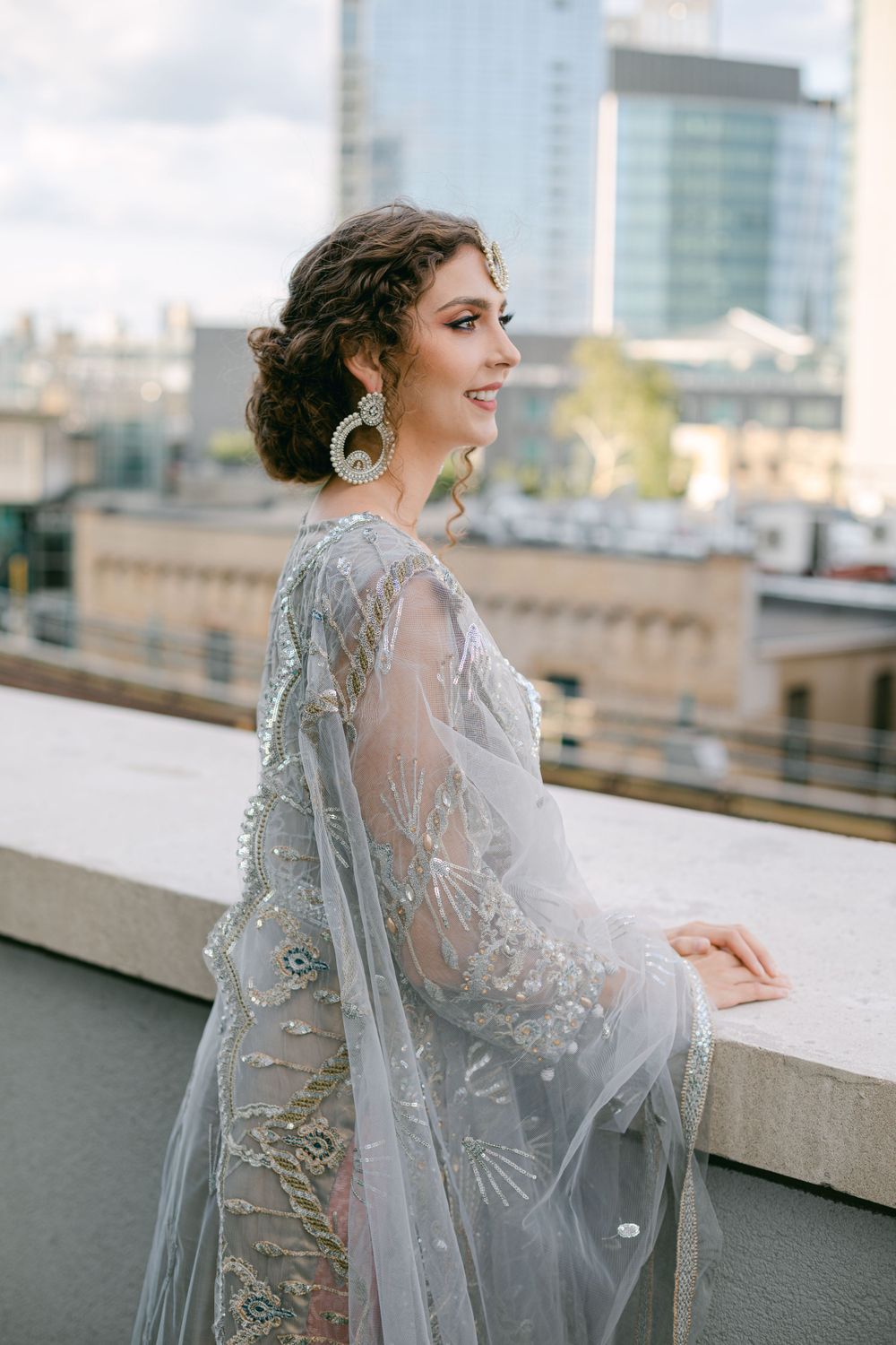 Elegant silver embellished sheer gown photographed on a rooftop with city buildings in the background.