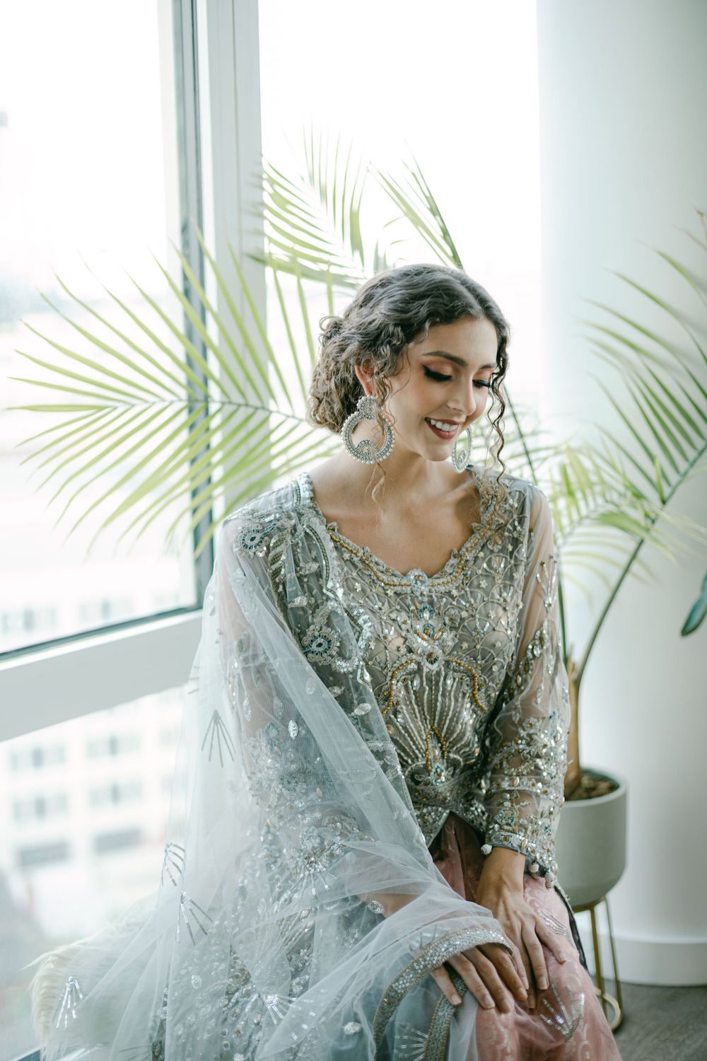 An elegant bridal portrait featuring a silver beaded gown and palm plants in a bright indoor setting.