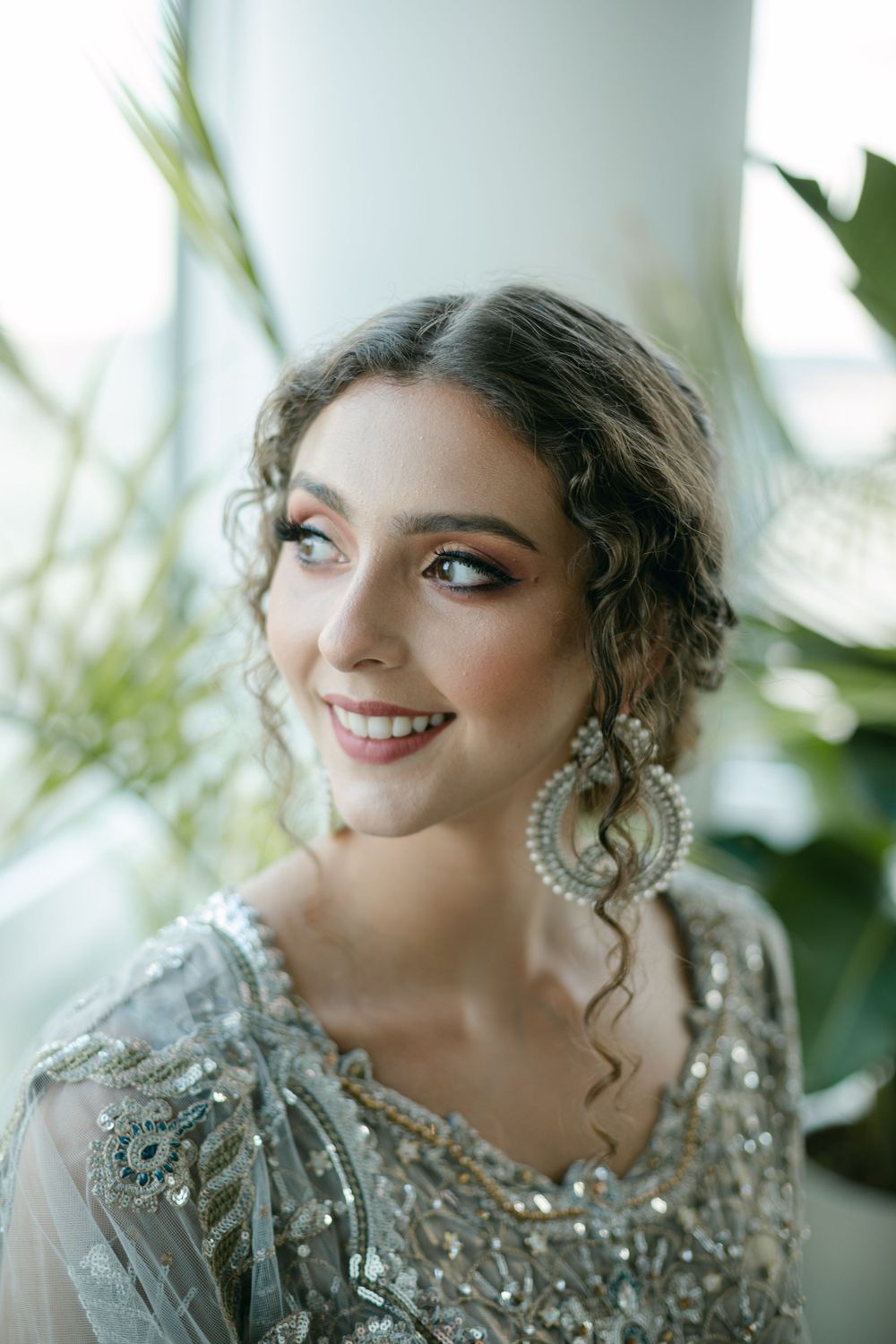 A bridal portrait with curled hair and statement earrings against a bright window backdrop.