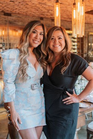 Two women pose together at an event, one wearing a light blue dress and one in a black dress with warm lighting.