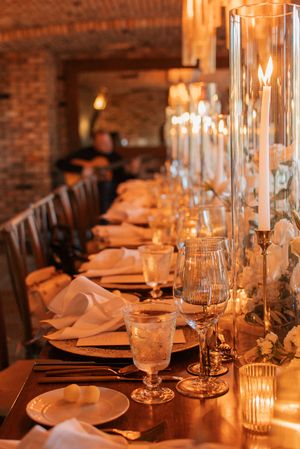 Candlelit dining table with elegant place settings, glassware, and brick wall backdrop.