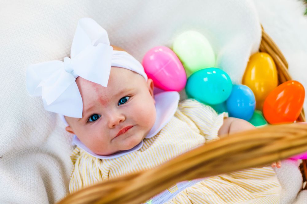 Baby wearing a white bow lies next to colorful plastic Easter eggs in a basket.